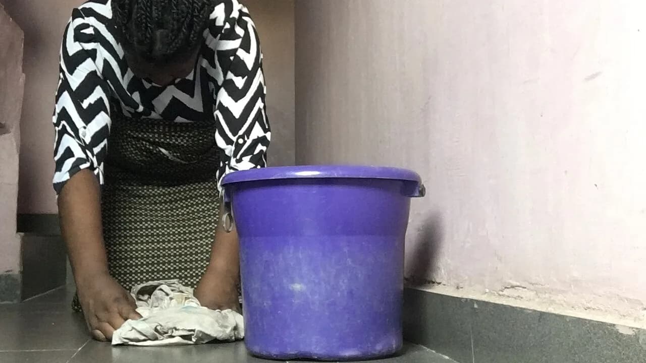 Seorang perempuan menyeka lantai keramik abu-abu dengan kain di lorong, di samping ember plastik