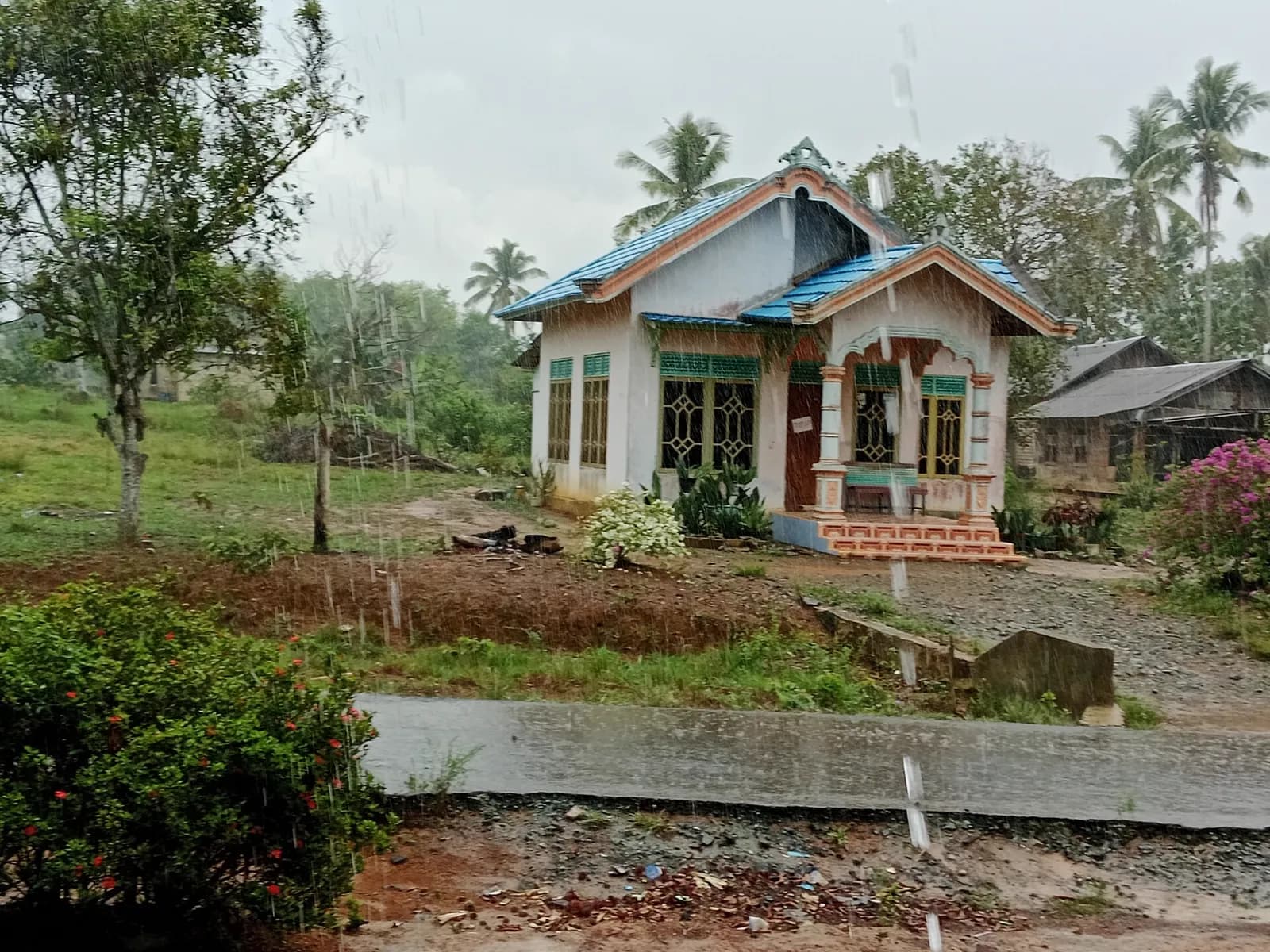 Rumah satu lantai di Indonesia saat hujan dengan jalan masuk kerikil melintasi parit lewat gorong-gorong beton, bukan foto proyek Inproland