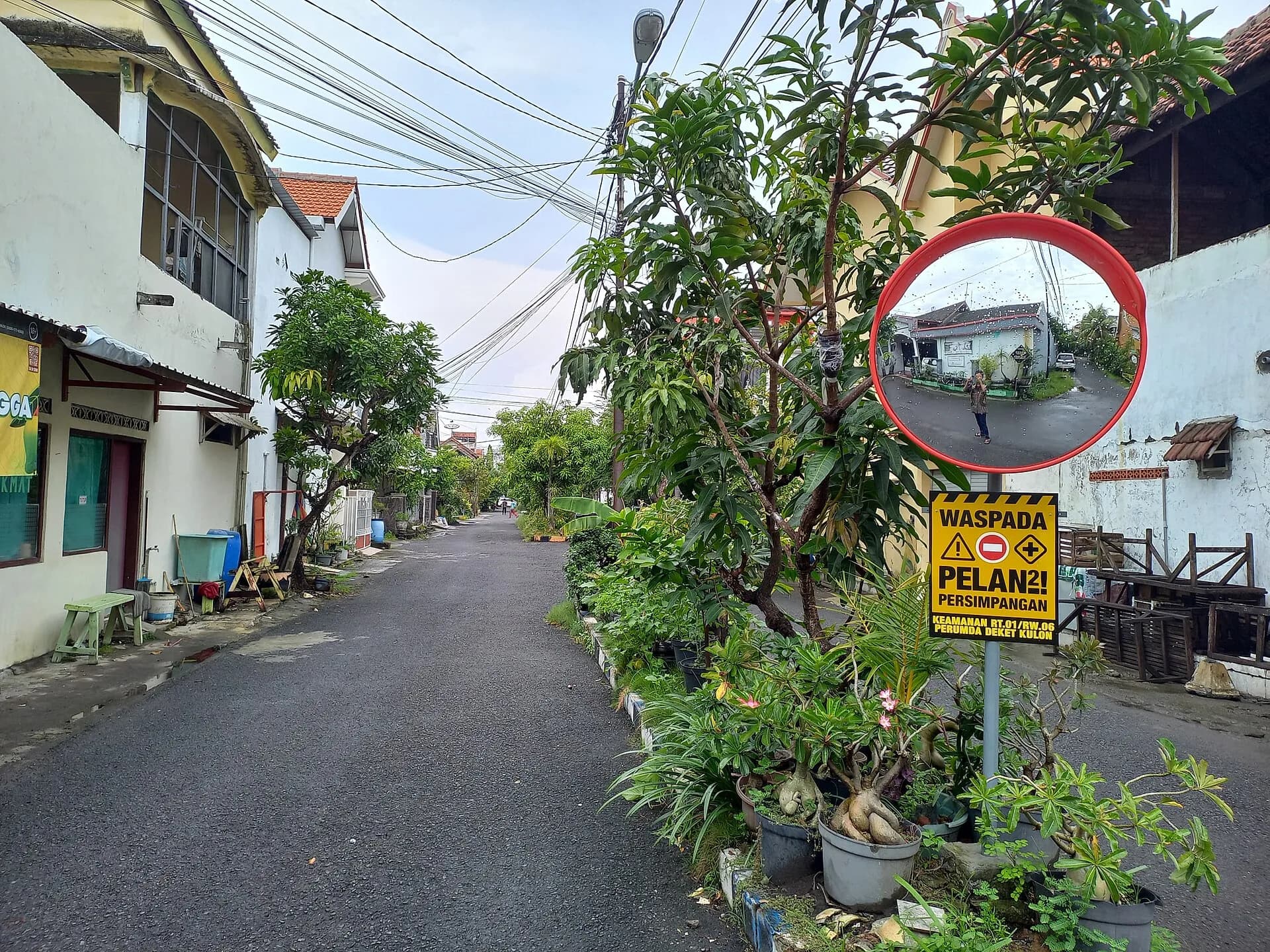 Jalan di lingkungan perumahan dengan deretan rumah, tanaman pot, dan cermin cembung di sudut jalan